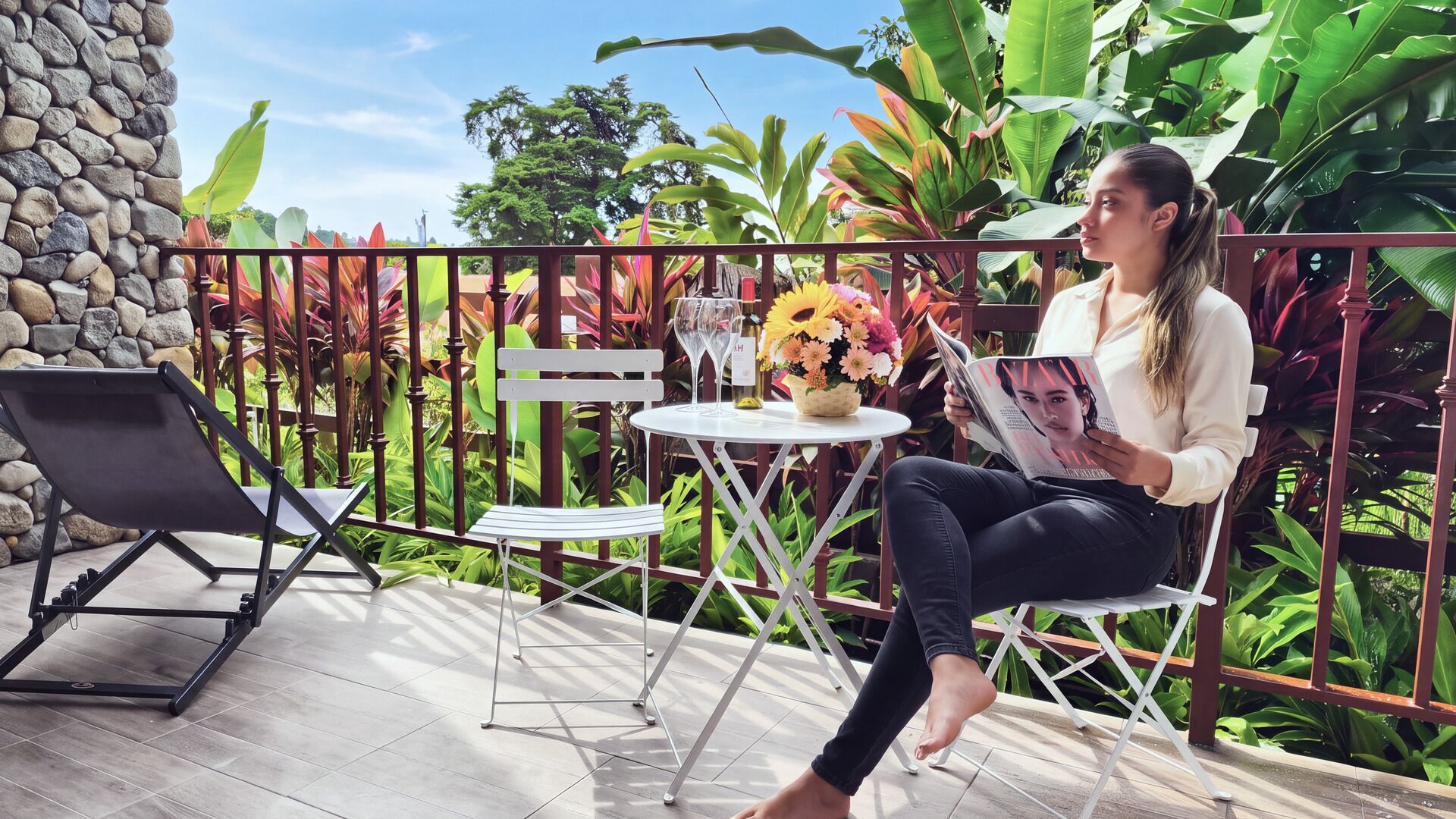 A person seated on a chair at a table with a drink and a flower arrangement, outdoors with a stone wall and tropical plants in the background.