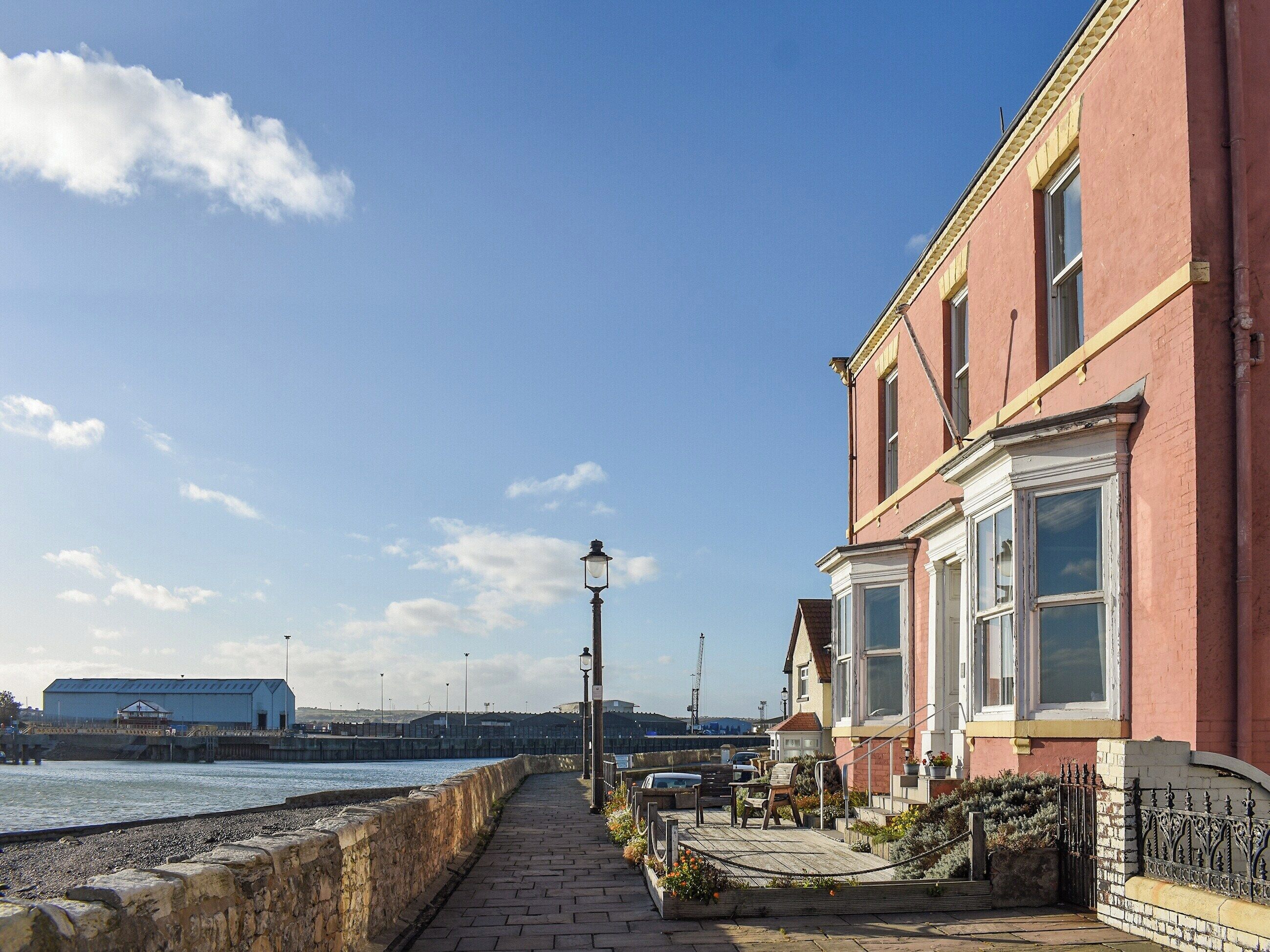 A waterfront promenade with a stone wall, a wooden deck, and a row of red brick buildings with large windows.