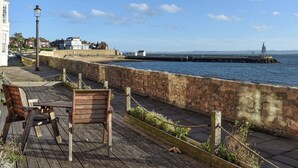 A coastal promenade with a wooden bench, stone wall, and a lighthouse in the distance.