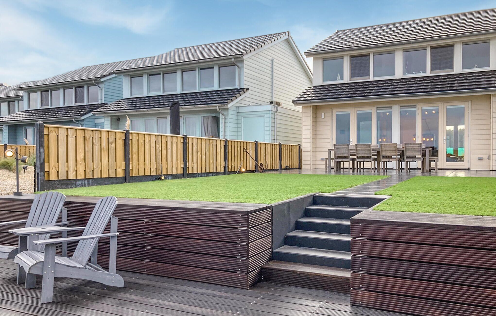 A residential property with a well-maintained lawn, wooden deck with chairs, and a staircase leading to a covered patio area.