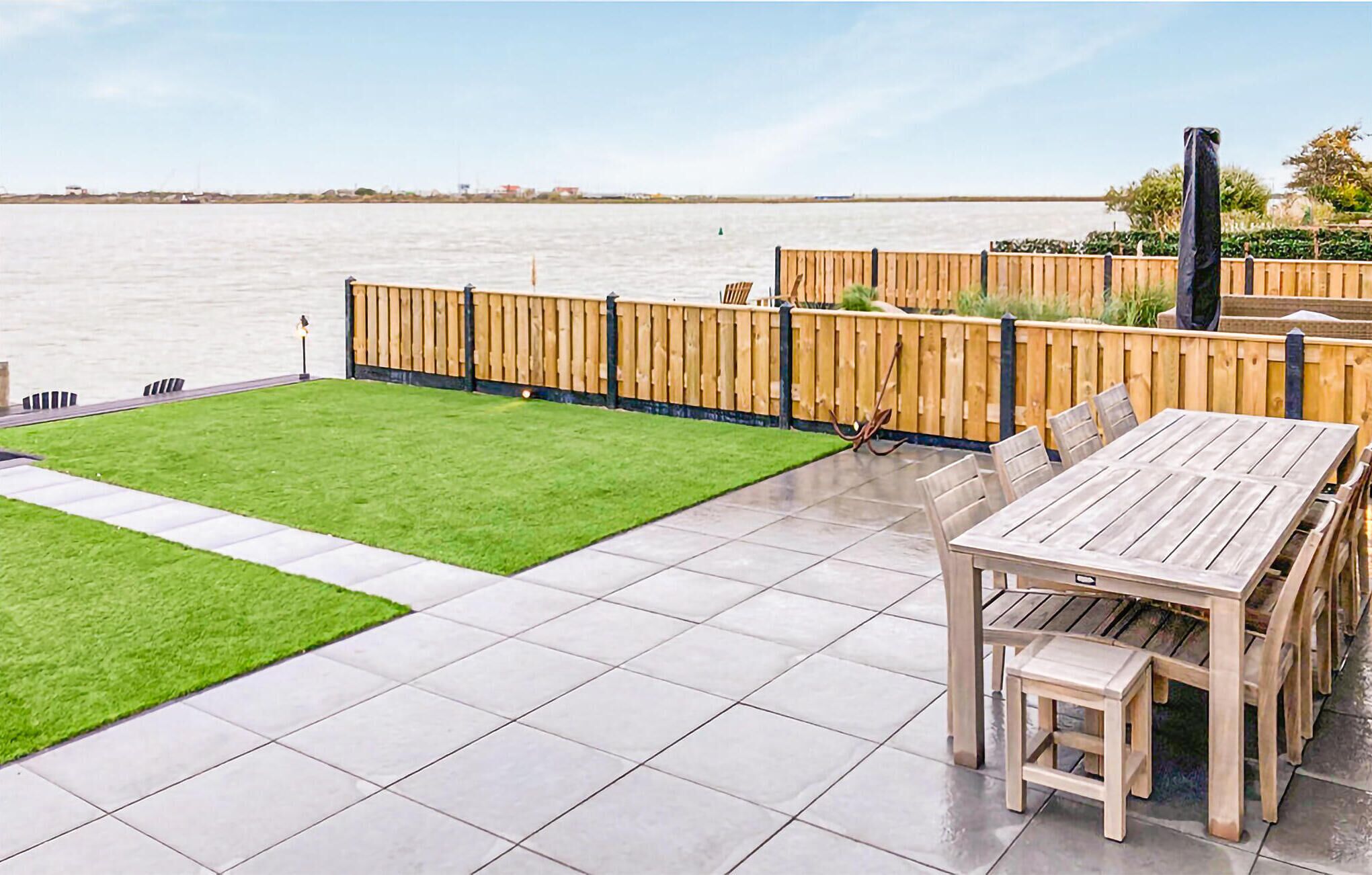 A patio with a wooden table and chairs, a small lawn, and a wooden fence overlooking a body of water.
