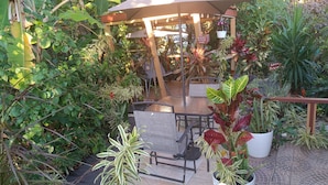 Un espacio de estar al aire libre con pérgola, mesas y sillas, rodeado de plantas en macetas y abundante vegetación.