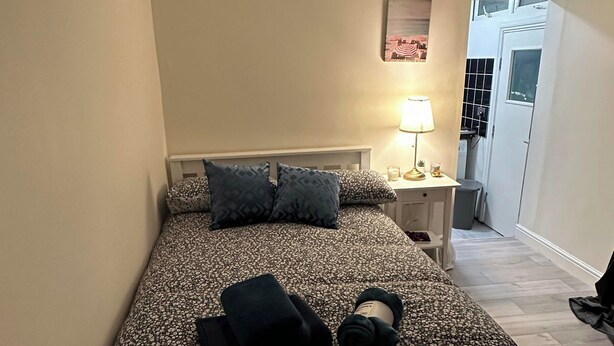 A bedroom with a bed, pillows, a bedside table with a lamp, a small table with a vase of flowers, and a door leading to another room. - Lovely Studio Apartment in London (London)