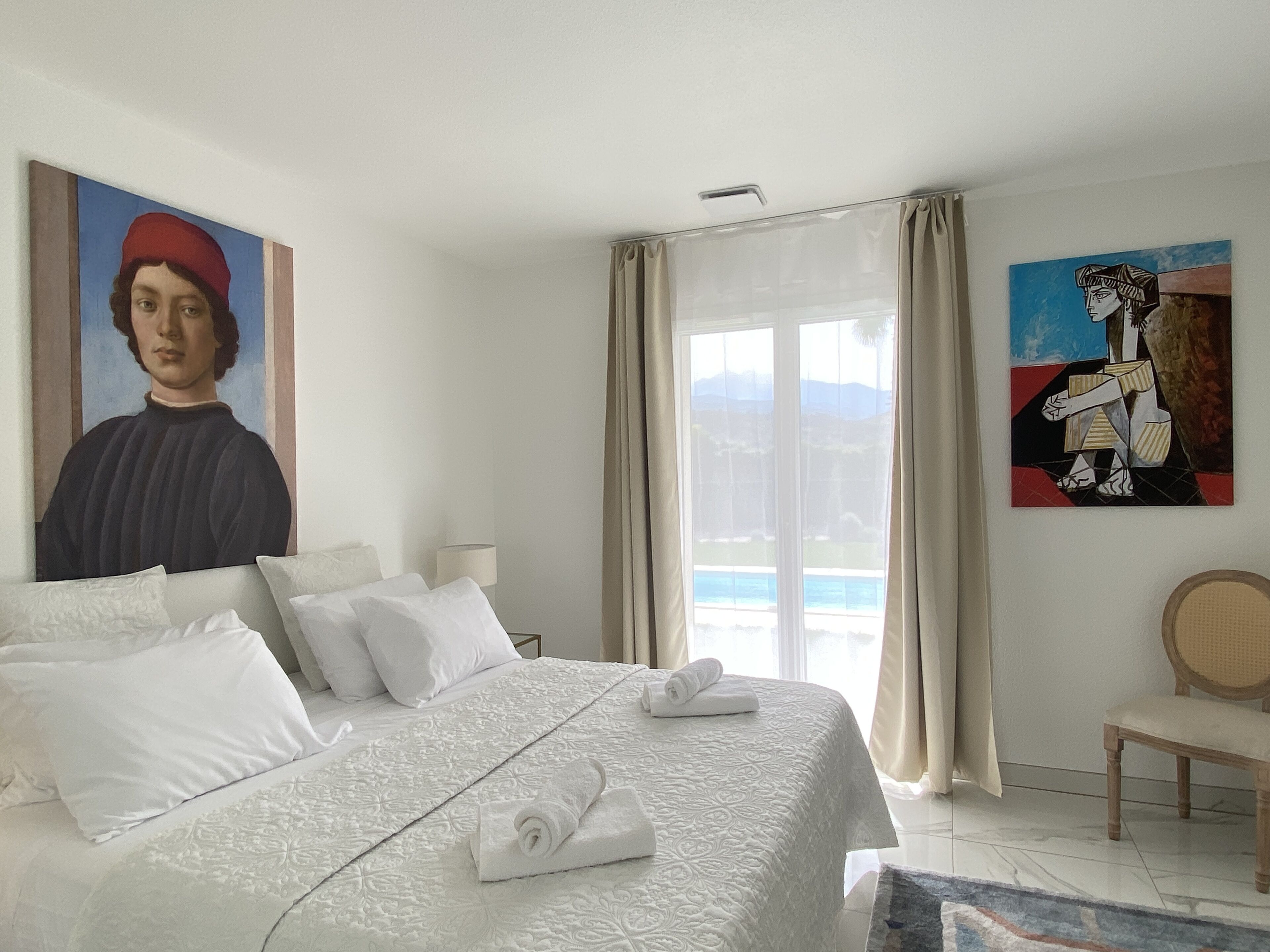 A hotel room with a bed, a chair, a painting, and a view of the sea.