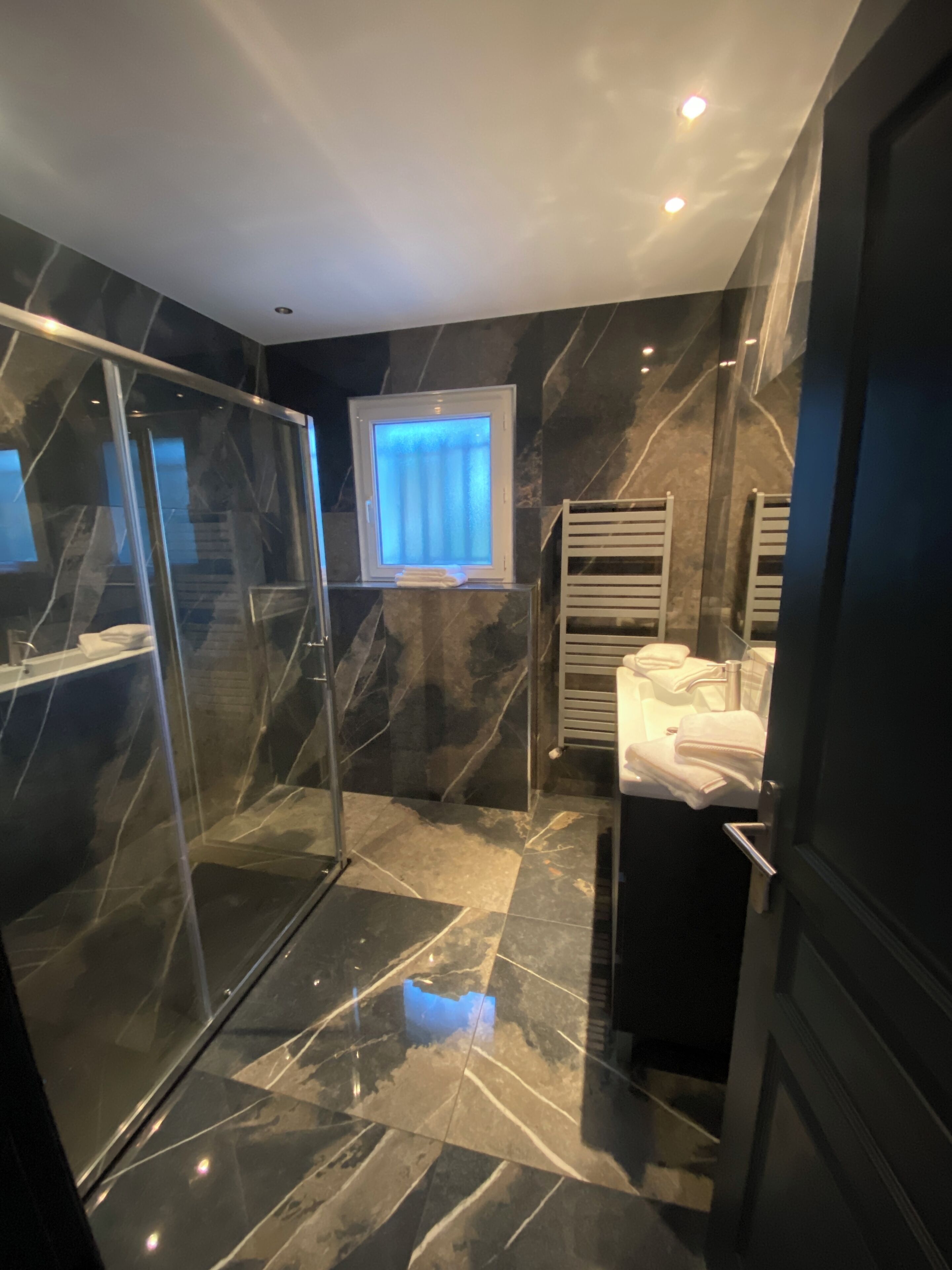 A modern bathroom with a glass shower enclosure, a window with curtains, a towel rack, and a sink with a mirror.