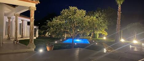 A pool area at night with a tree, lounge chairs, and a building with columns.