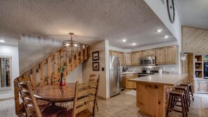 Eine geräumige Küche mit Holzschränken, einer zentralen Kochinsel mit Barhocker sowie einem Edelstahlkühlschrank und einer Holztreppe.