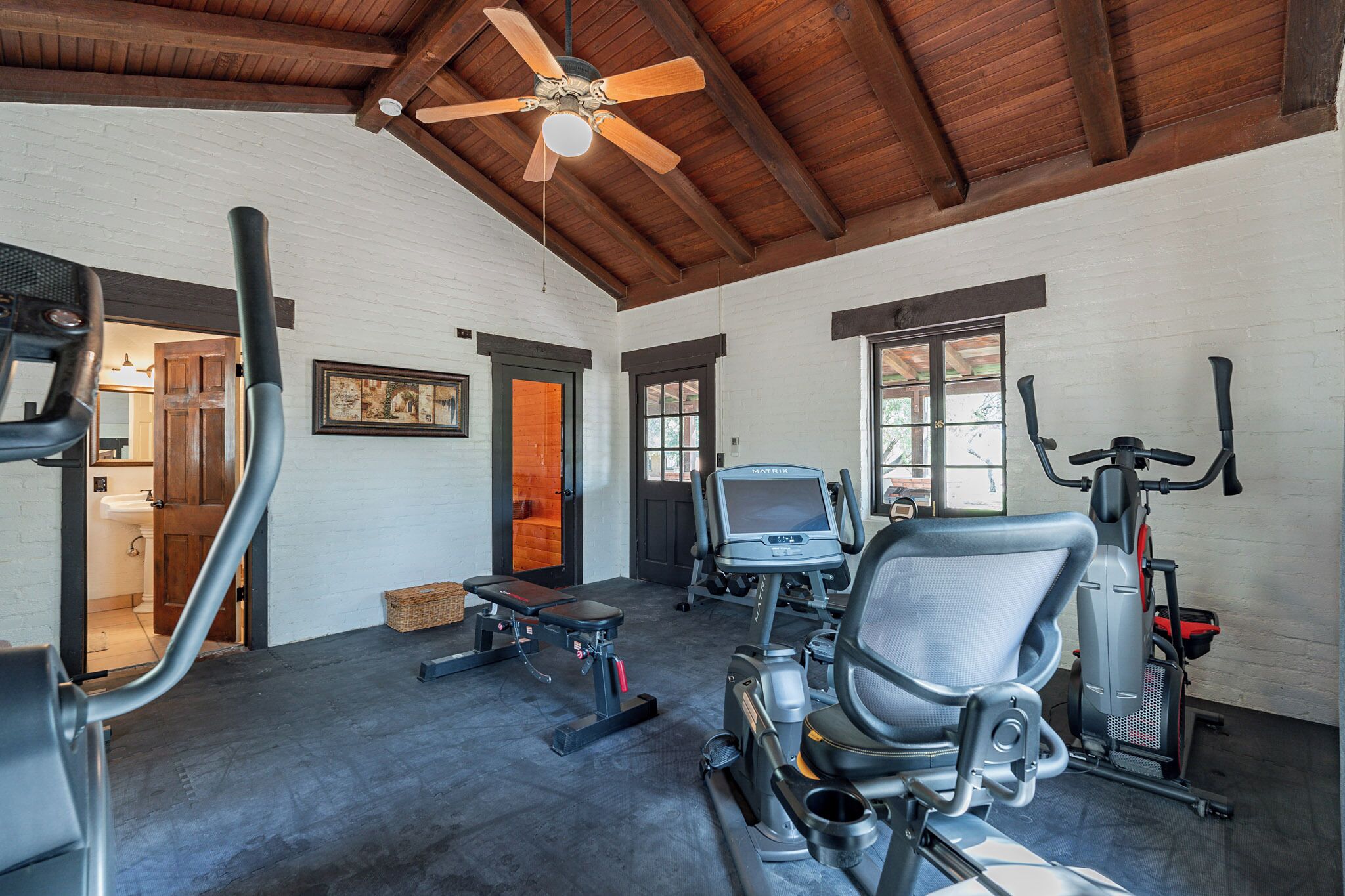 Ein Fitnessraum mit Trainingsgeräten, einem Deckenventilator und Blick ins Freie.