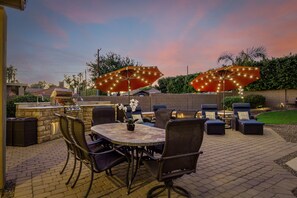 An outdoor patio with a stone table, wicker chairs, and two large umbrellas with string lights. - Heated pool, jacuzzi, Fast Wifi, Pac Man, Fire Pit, BBQ, Sleeps 14! (Phoenix)