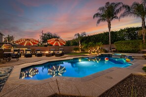 A swimming pool with a fire pit and outdoor seating area, surrounded by palm trees and a sunset sky. - Heated pool, jacuzzi, Fast Wifi, Pac Man, Fire Pit, BBQ, Sleeps 14! (Phoenix)