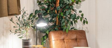 Una habitación con un sillón de cuero marrón, una mesita redonda con una lámpara, una planta en maceta y un cuadro en la pared.