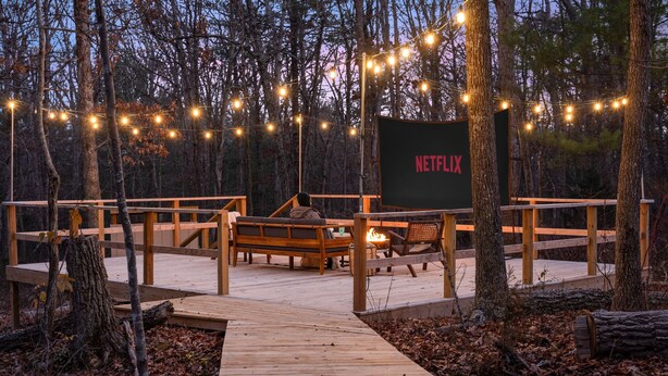 A wooden deck with a bench and a fire pit, surrounded by trees and string lights. - Outdoor Theatre | Hot Tub | Fire Pit | Dogs (Luray)