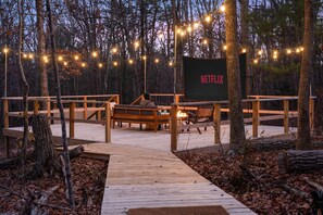 A wooden deck with a bench and a fire pit, surrounded by trees and string lights. - Outdoor Theatre | Hot Tub | Fire Pit | Dogs (Luray)
