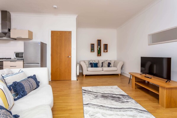 A modern living room with a sofa, TV, and a kitchen area in the background. - No27 The Barclay - Donnini Apartments (Kilmarnock)