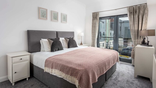 A bedroom with a bed, nightstand, and a view of the outside through a glass door. - No27 The Barclay - Donnini Apartments (Kilmarnock)