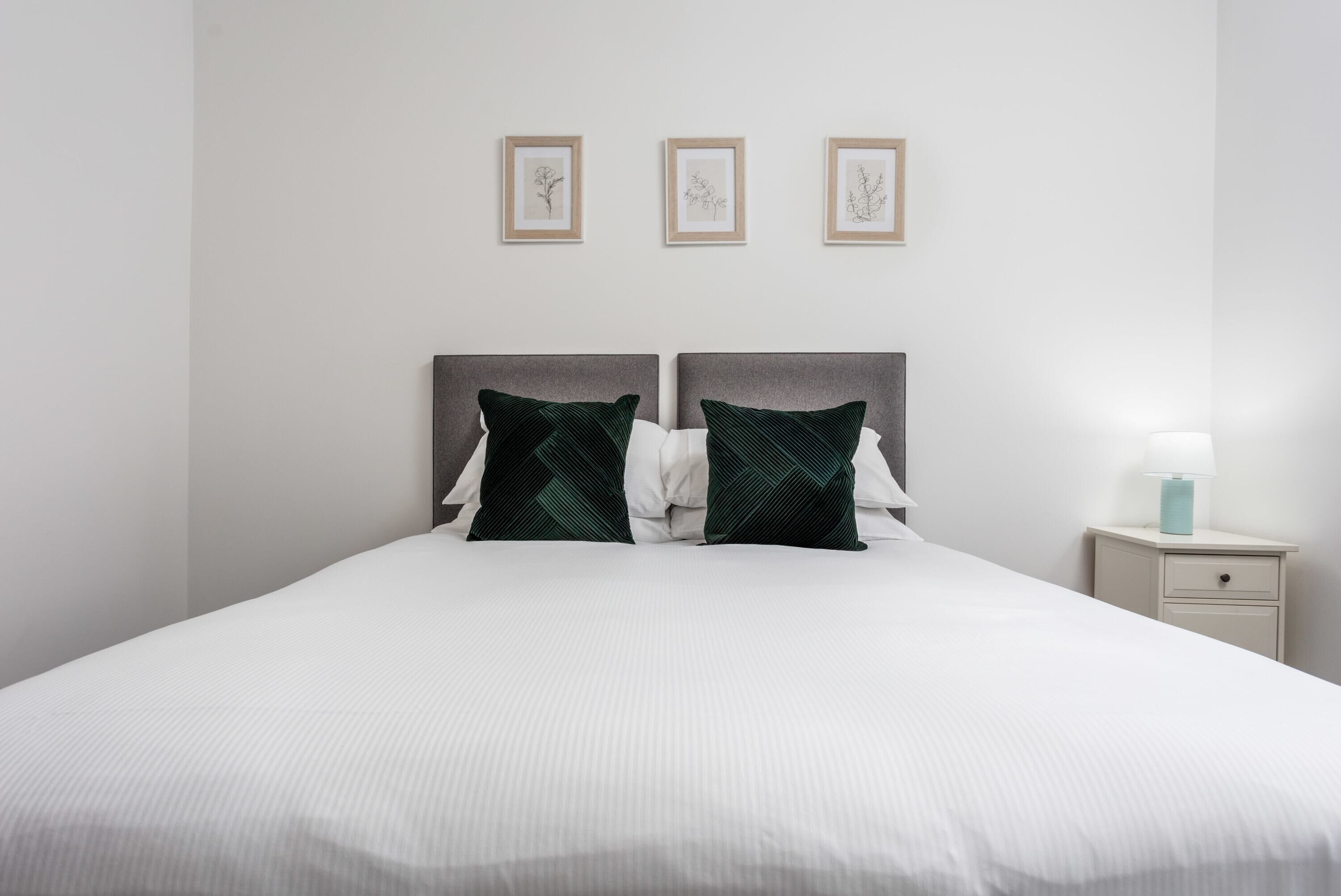 A neatly made bed with two pillows, a bedside table with a lamp, and three framed botanical prints on the wall.