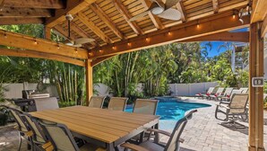An outdoor patio with a pool, wooden table, chairs, and a grill. - Beach Bound: 1 Block to Beach! Pool & Spa! Perfect AMI location! by Anna Maria Life Vacation Rentals (Holmes Beach)