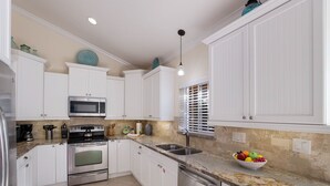 A modern kitchen with white cabinetry, stainless steel appliances, a granite countertop, and a bowl of fruit. - Beach Bound: 1 Block to Beach! Pool & Spa! Perfect AMI location! by Anna Maria Life Vacation Rentals (Holmes Beach)