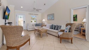 A spacious living room with a high ceiling, a ceiling fan, and a large window. There is a sofa, two wicker chairs, and a coffee table. - Beach Bound: 1 Block to Beach! Pool & Spa! Perfect AMI location! by Anna Maria Life Vacation Rentals (Holmes Beach)