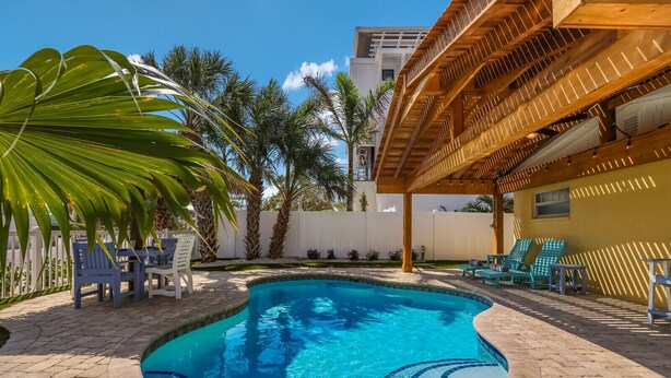 A swimming pool with a wooden pergola, palm trees, and lounge chairs.