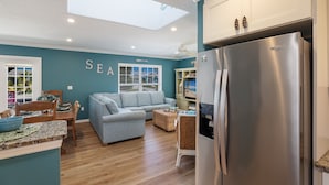 A modern kitchen with a stainless steel refrigerator, a dining area with a wooden table and chairs, a light blue sofa, and a ceiling skylight.