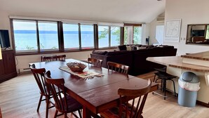 Een eetgedeelte met een houten tafel en stoelen, een donkerbruine bank en uitzicht op zee door grote ramen.