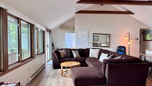 Een woonkamer met een bruine modulaire bank, een ronde houten salontafel en een groot raam met uitzicht op bomen.