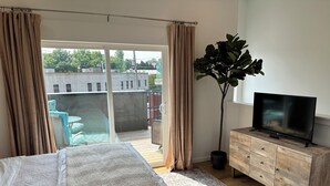 Un dormitorio con cama, televisor sobre un mueble de madera, planta en maceta y vistas al balcón exterior.