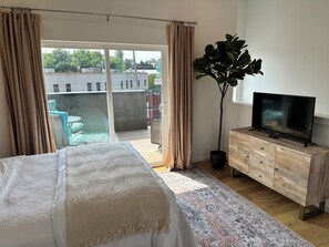 Une chambre à coucher avec un lit, un téléviseur posé sur un meuble en bois, une plante en pot et une vue sur le balcon extérieur.