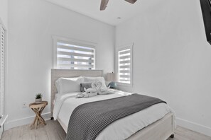 Une chambre à coucher avec un lit, une petite table de chevet en bois sur laquelle est posée une plante, et deux fenêtres équipées de stores.