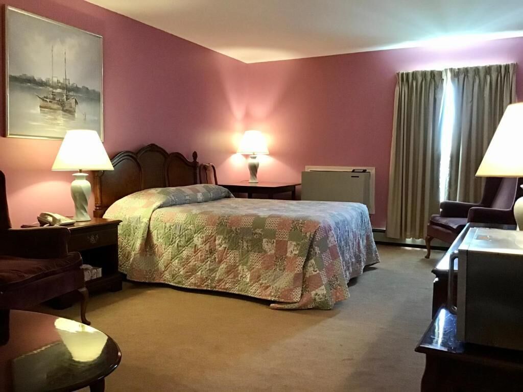 Quarto de hotel com cama, duas luminárias de cabeceira, escrivaninha, cadeira, mesa de cabeceira, luminária, um quadro na parede e uma janela com cortinas.