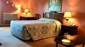 A hotel room with a bed, two bedside lamps, a nightstand with a lamp, a chair, a table, a painting on the wall, and a radiator.