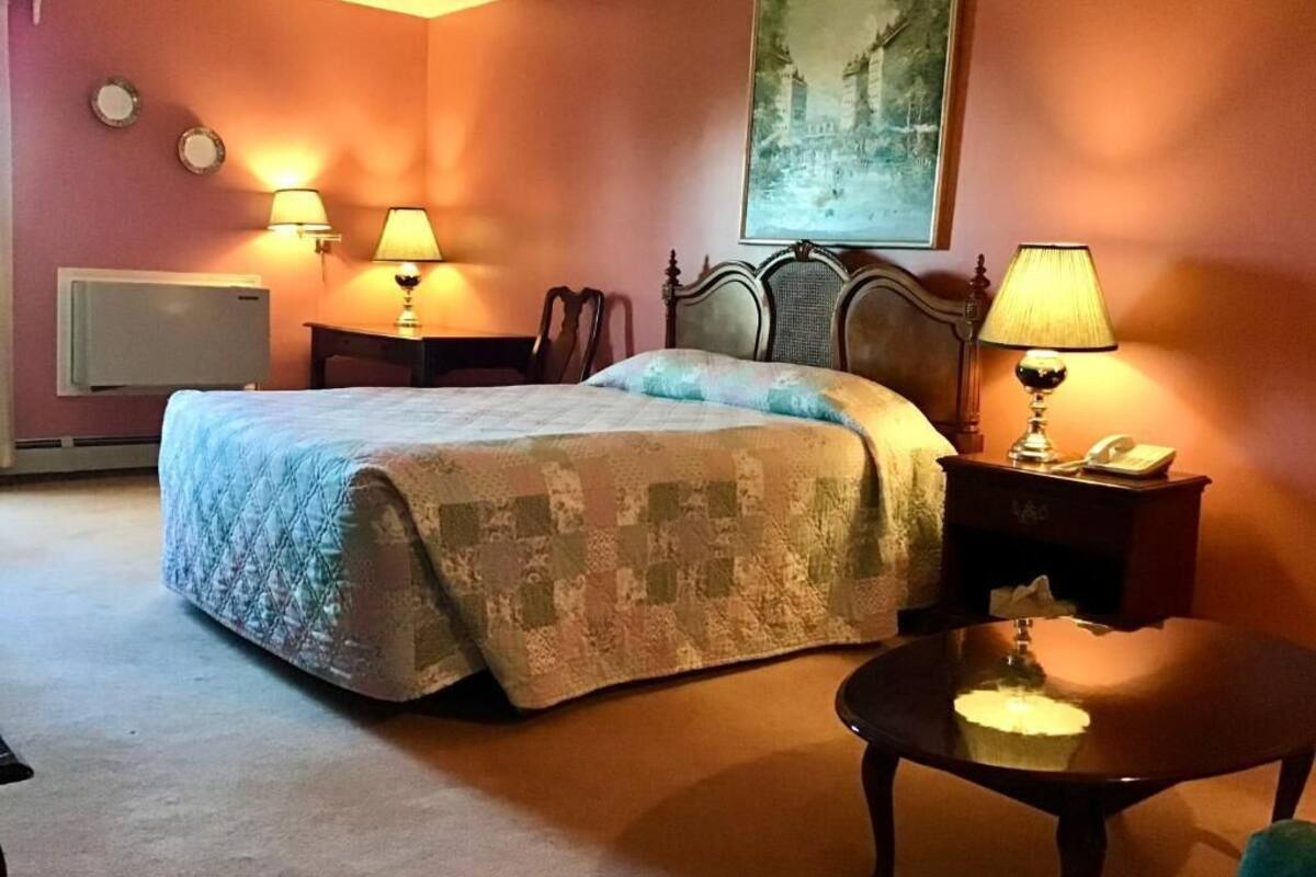 A hotel room with a bed, two bedside lamps, a nightstand with a lamp, a chair, a table, a painting on the wall, and a radiator.