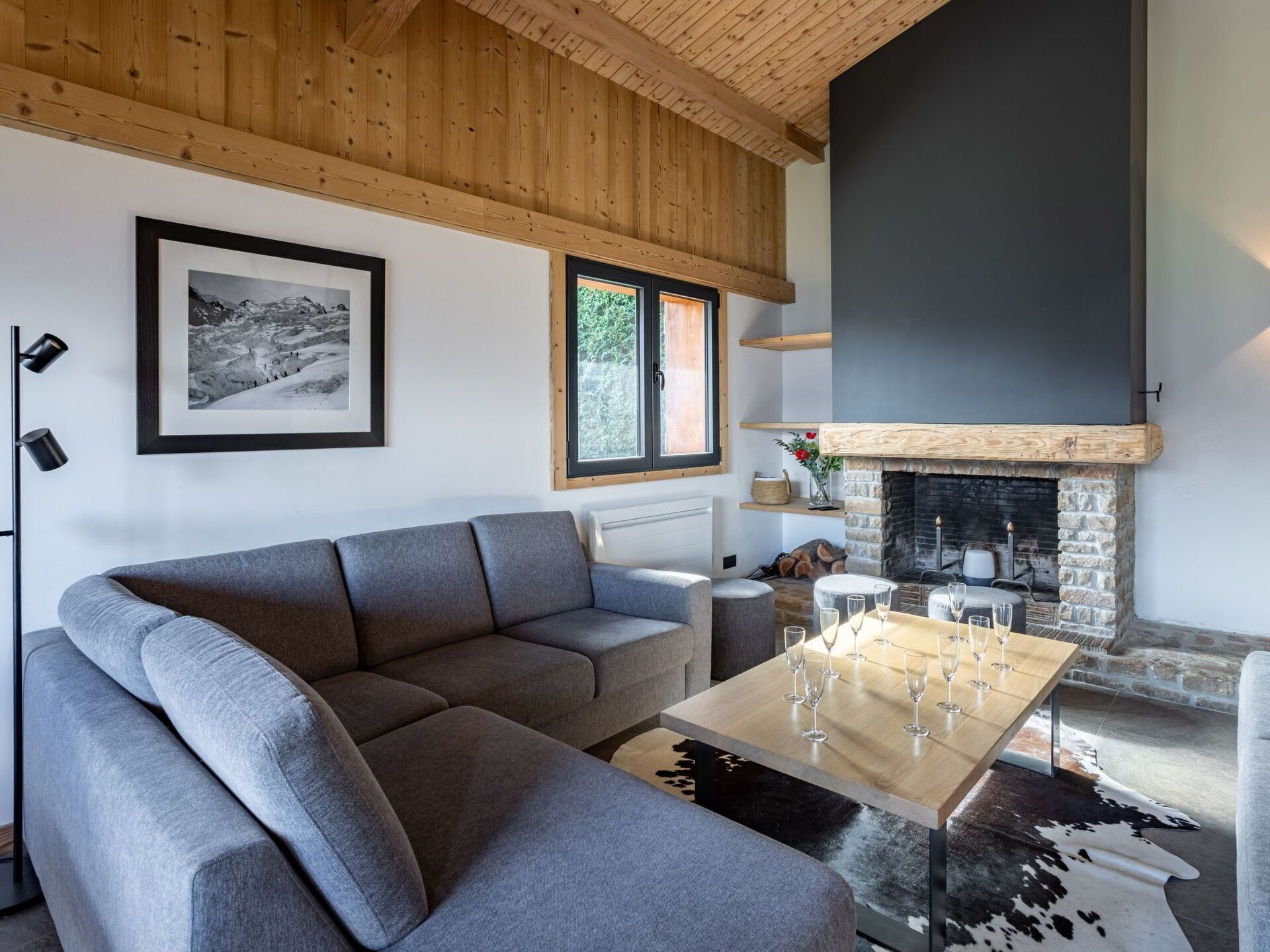 A modern living room with a grey sectional sofa, a wooden coffee table with wine glasses, a stone fireplace, and a framed photograph on the wall.