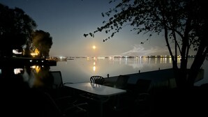Een terras aan het meer met een tafel en stoelen, uitzicht op de maan, en een rustig waterlichaam dat lichtjes weerspiegelt.
