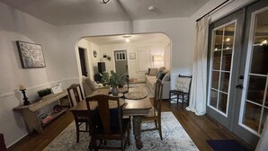 A dining area with a table set for four, surrounded by chairs. There is a large window with curtains, a door leading to another room, and a wall-mounted shelf with decorative items.