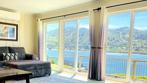 A spacious living room with a large window offering a scenic view of a lake and mountains. - River’s End-Best Pacific Ocean and Rogue River Views on 2+Acres with a Hot Tub!  (Gold Beach)