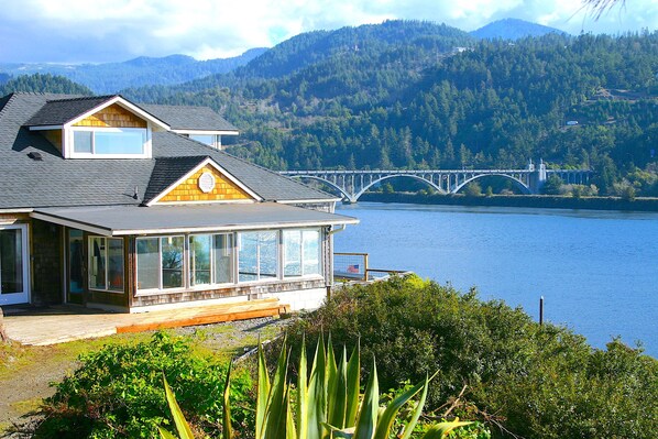 A house with a large bay window overlooking a lake and a bridge. - River’s End-Best Pacific Ocean and Rogue River Views on 2+Acres with a Hot Tub!  (Gold Beach)