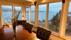 A dining area with a wooden table, chairs, and a vase of flowers. Large windows offer a view of the sea and distant land. - River’s End-Best Pacific Ocean and Rogue River Views on 2+Acres with a Hot Tub!  (Gold Beach)