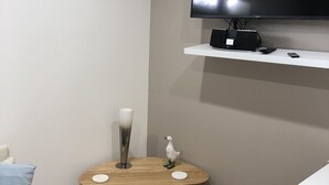 A modern living room with a flat-screen TV mounted on the wall, a small wooden side table with a decorative item, and a white couch with a blue pillow.