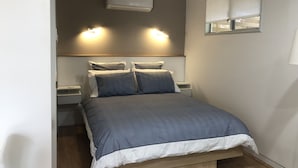 A modern bedroom with a bed, bedside tables, an air conditioning unit, and a window with blinds.