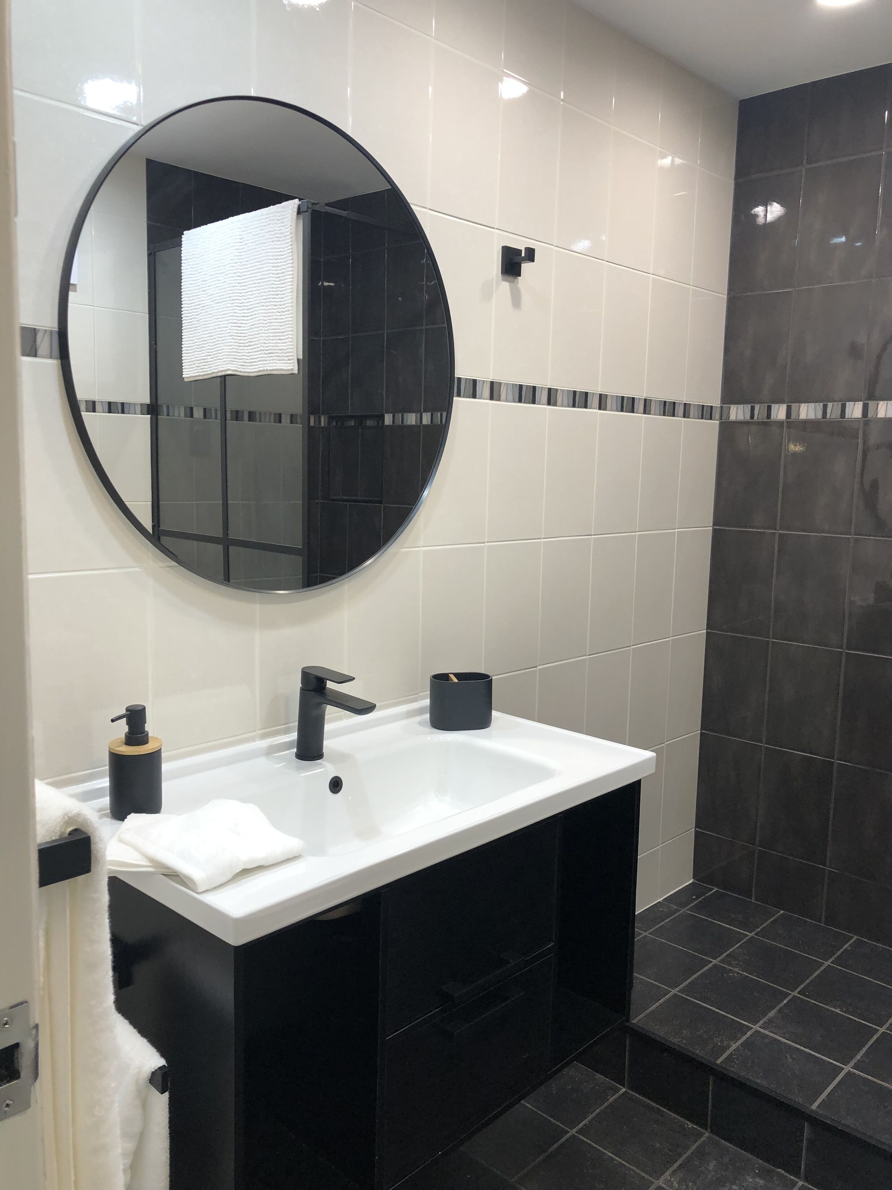 A modern bathroom with a round mirror, a white sink, black cabinets, and a shower area.