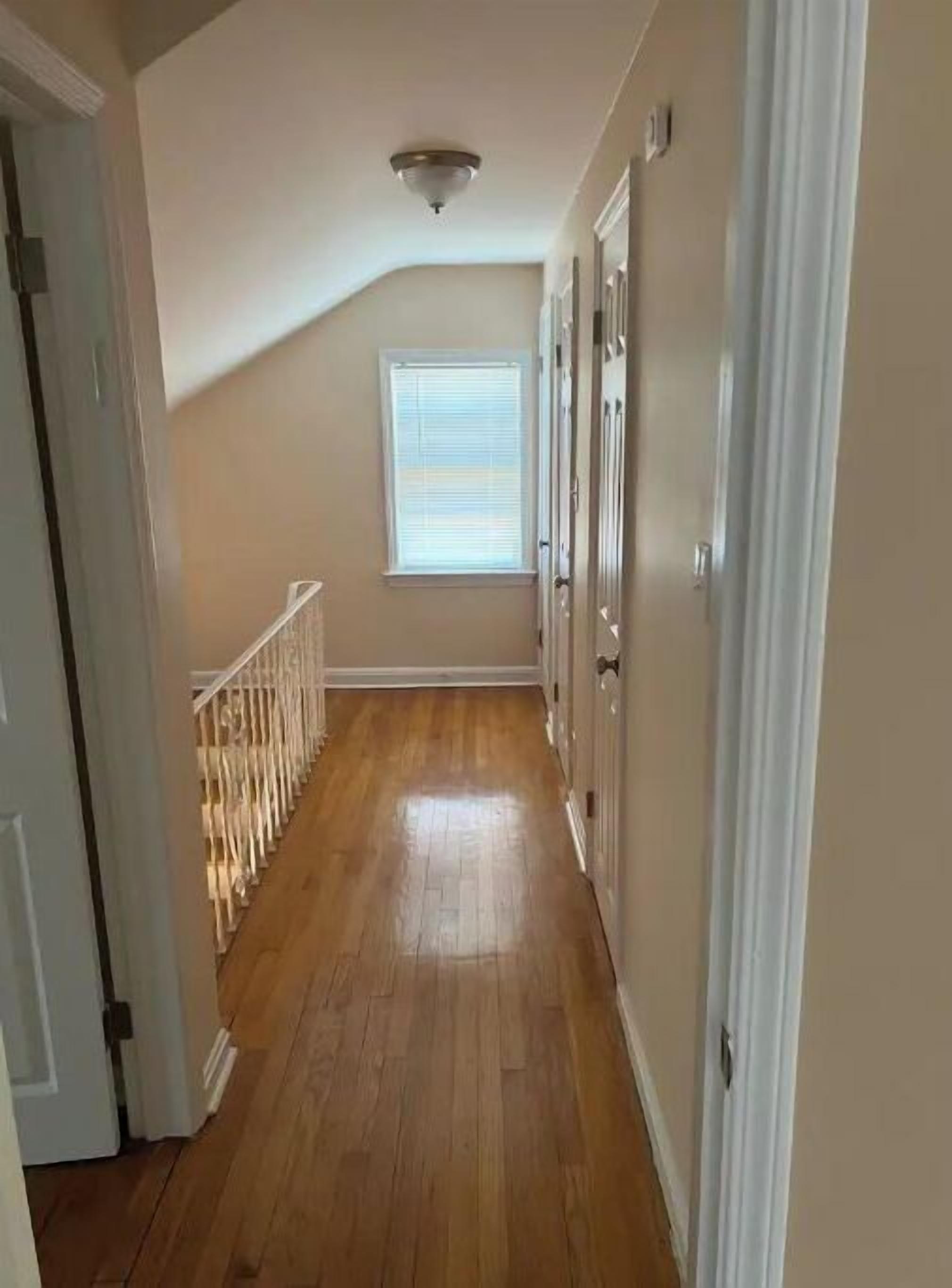 A narrow hallway with wooden flooring, a window with blinds, and a door on the left.