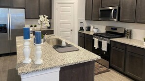 A modern kitchen with dark cabinets, granite countertops, a stainless steel refrigerator, a microwave, a stove, and a sink. - 4 BR Cheerful Home Long Term Stay Equipped in Katy (Katy)