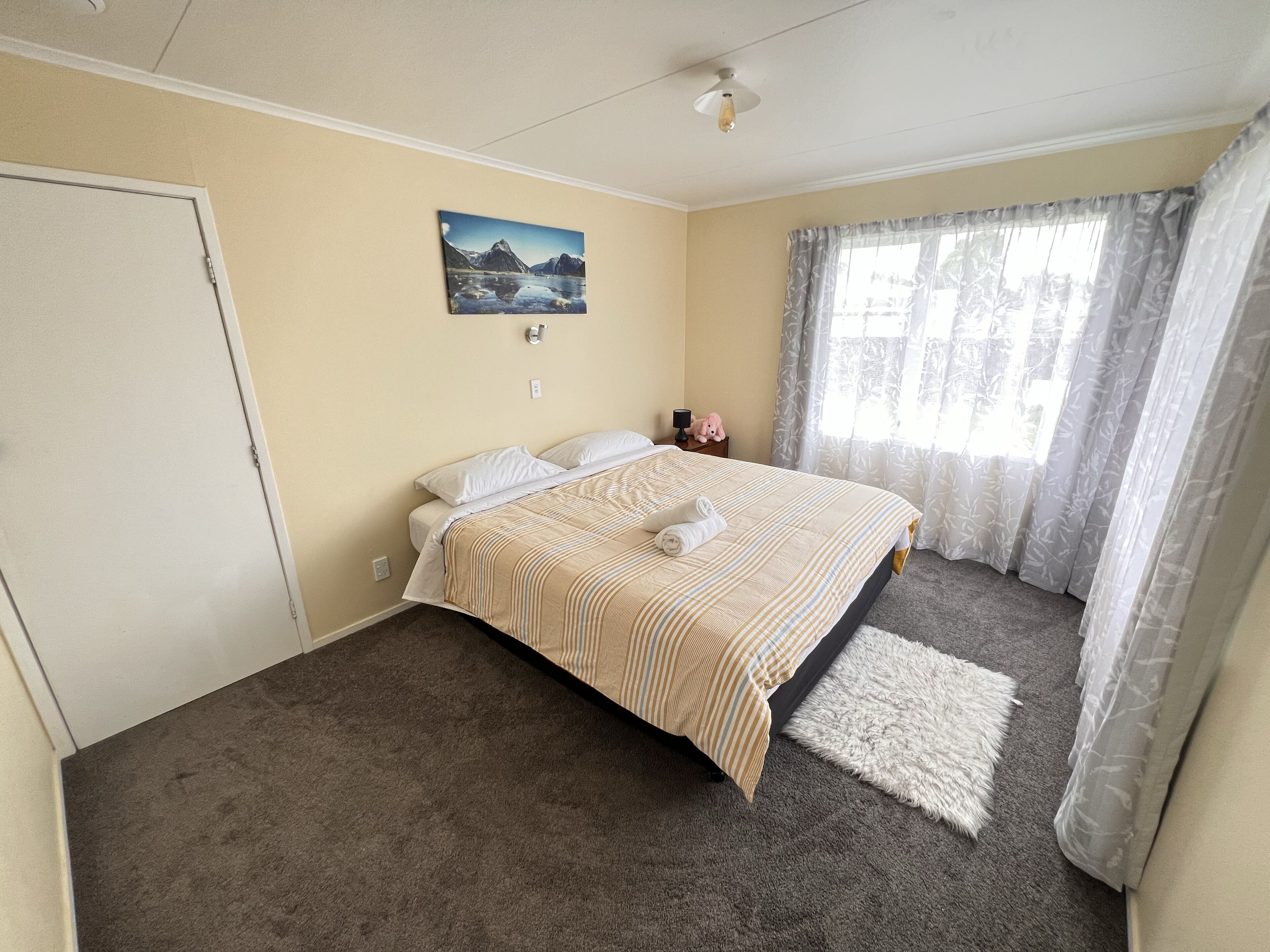 A bedroom with a bed, a painting, a nightstand, a lamp, and a window with curtains.