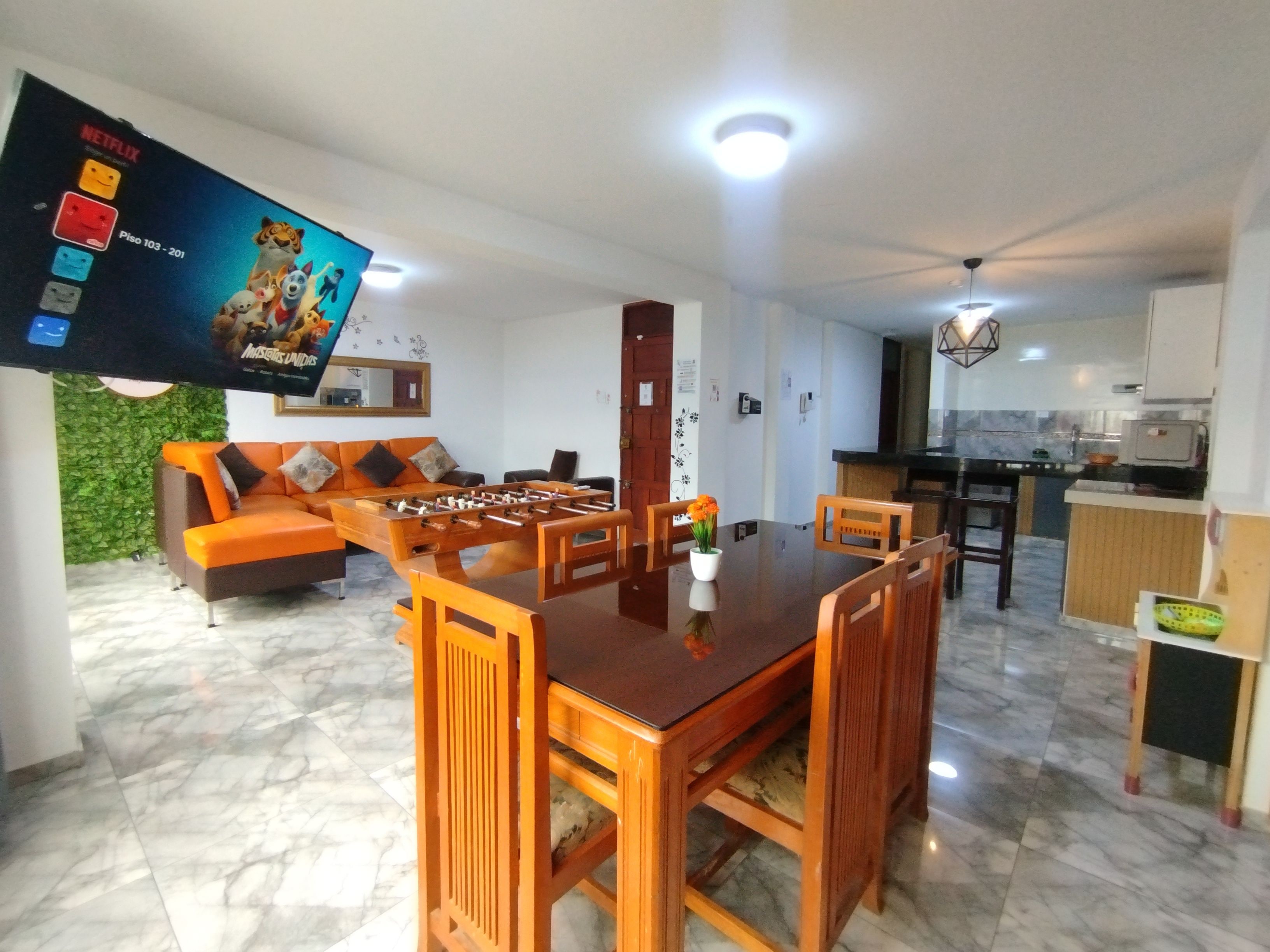 Una sala de estar con una mesa de fútbol, un sofá, un televisor montado en la pared y una zona de cocina al fondo.