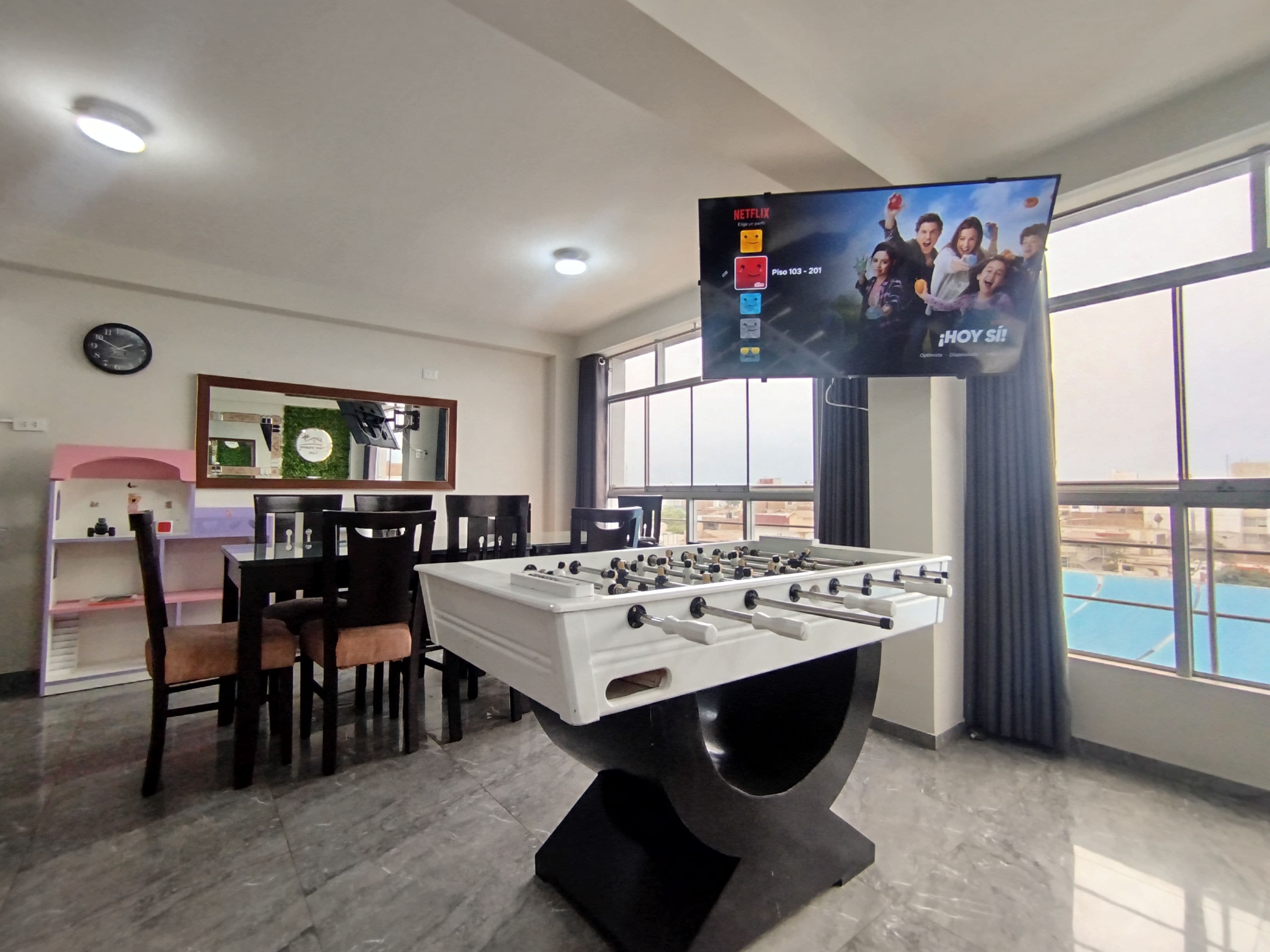 Una habitación con mesa de fútbol, televisor montado en la pared, un reloj y ventana con vistas.
