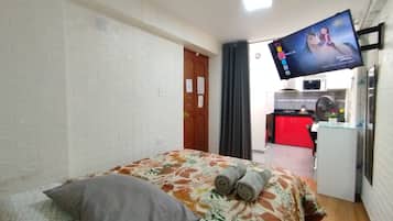 Habitación de hotel con cama, televisor de pantalla plana en la pared, kitchenette al fondo y puerta que da a otra habitación.