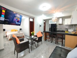 A modern living room with a flat-screen TV, a dining area with a kitchen in the background, and a bed with a white headboard.