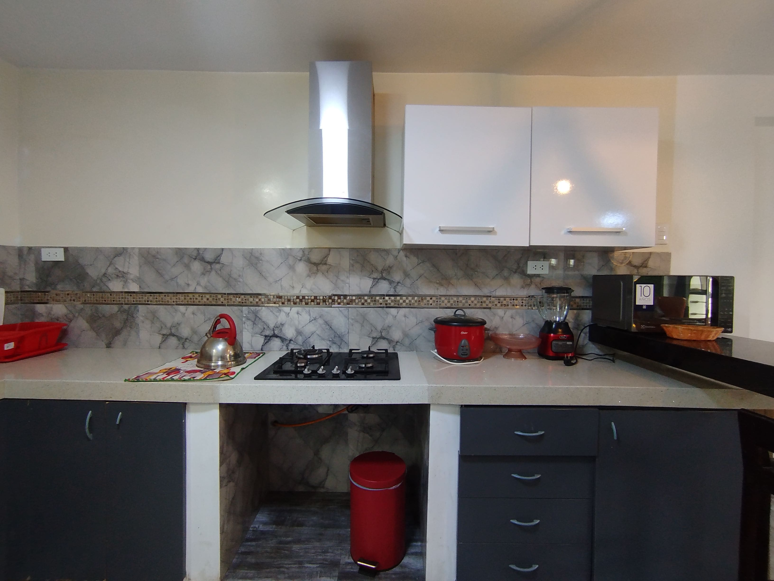Una cocina con estufa a gas, hervidor rojo, bote de basura rojo y microondas.